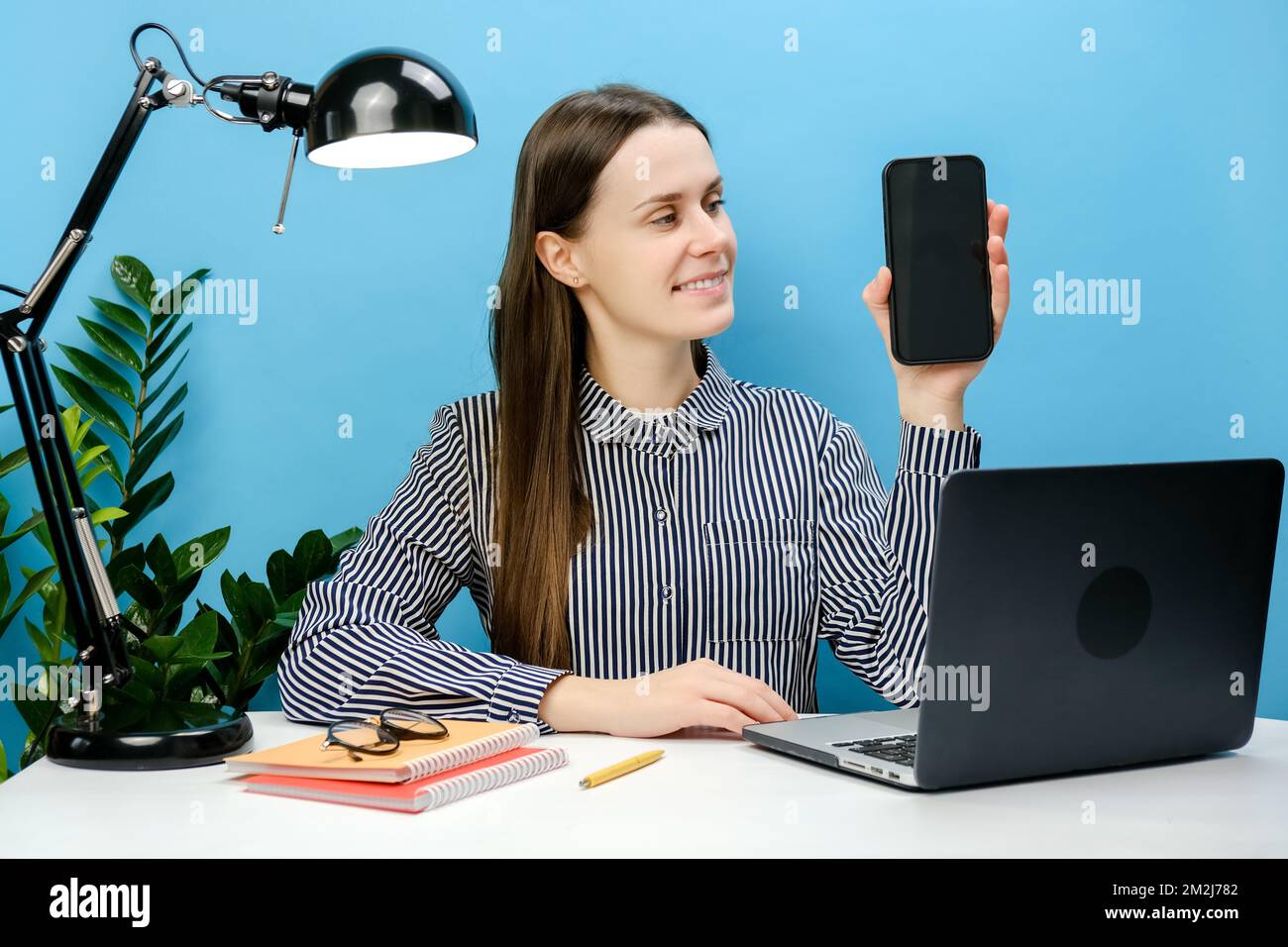 Felice donna di successo del lavoro del dipendente in camicia seduta lavoro alla scrivania bianca ufficio con laptop che tiene il telefono cellulare schermo vuoto area di lavoro, Foto Stock