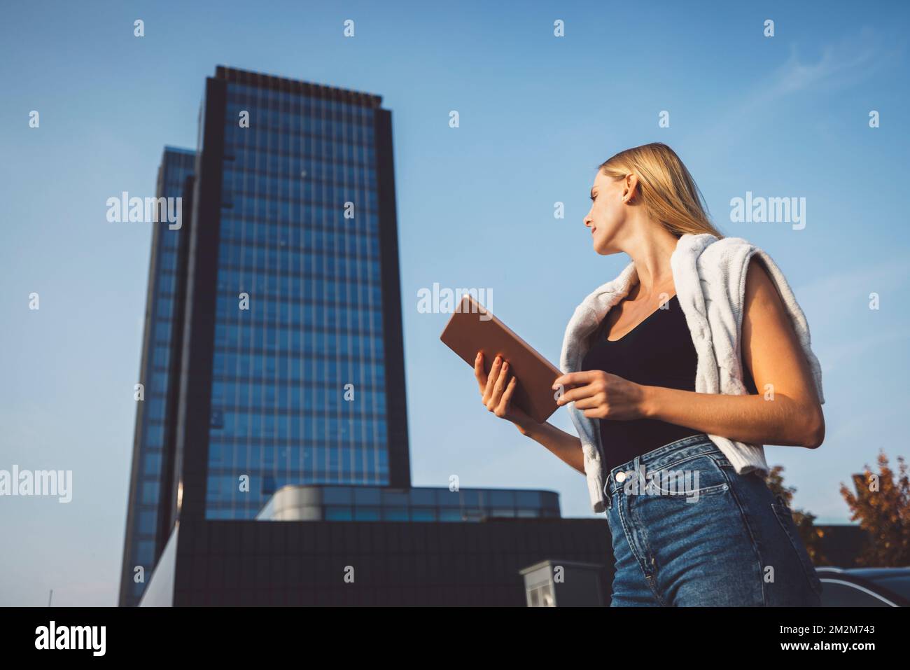 Vista dal basso angolo della donna bionda che tiene un tablet digitale con grattacielo sullo sfondo Foto Stock