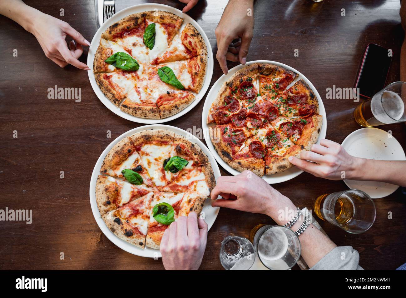 Vista dall'alto tre deliziose pizze italiane sul tavolo. Mani di persone tagliate irriconoscibili che prendono una fetta. Foto Stock