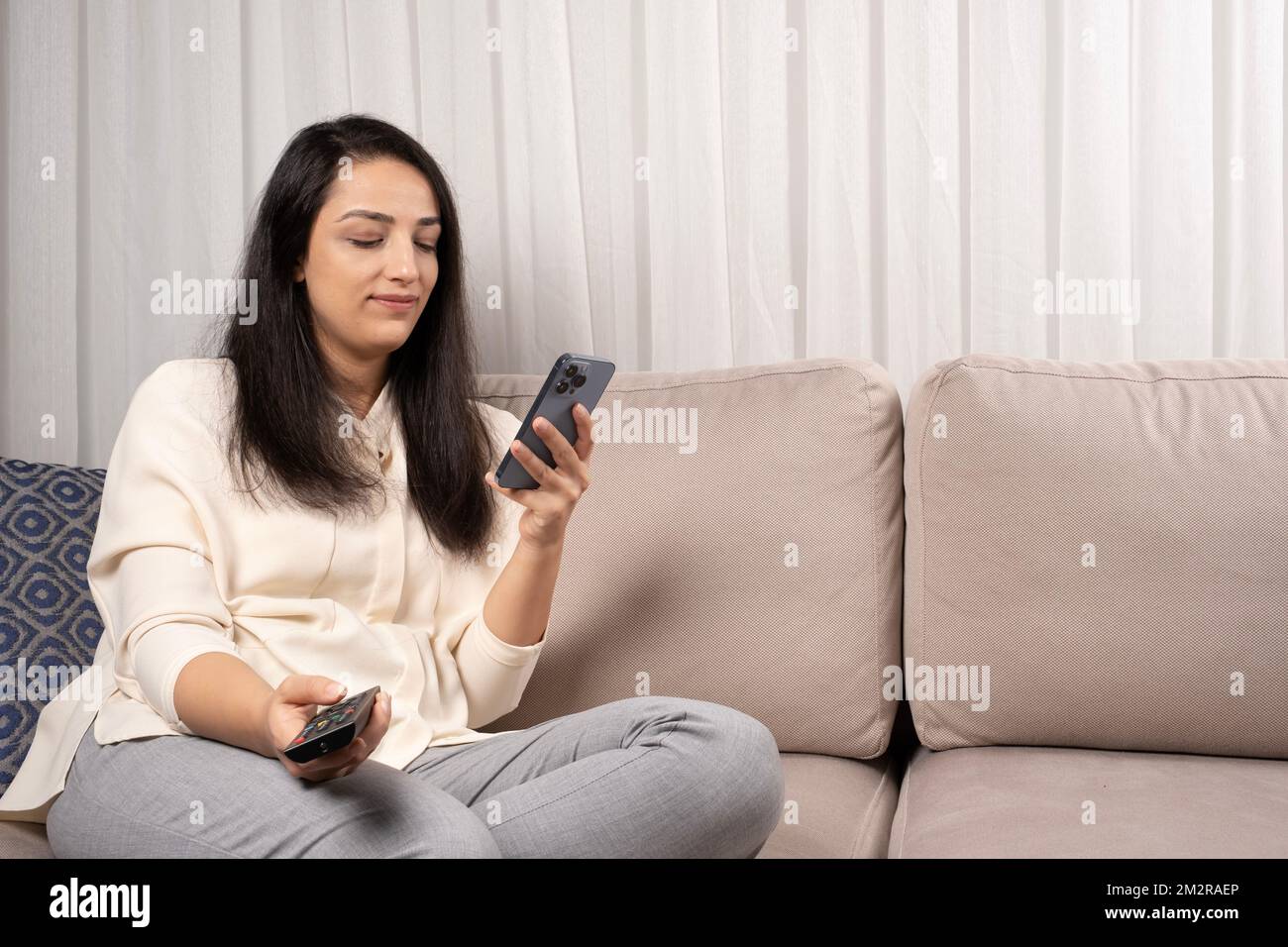 Ritratto di donna caucasica seduta sul divano con telecomando tv guardare smartphone. Internet, telefono o social media dipendenza idea foto. Foto Stock