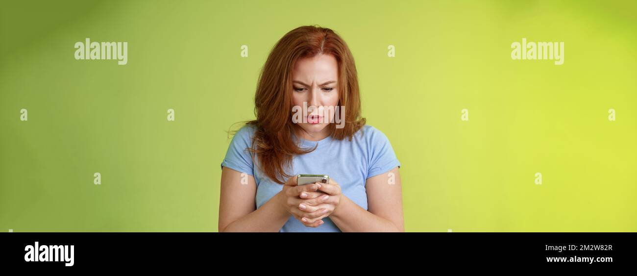 Confuso e incerto rosso donna di mezza età imparare come utilizzare i social media provare a capire emoji aspetto intenso e mirato smartphone display lettura importante Foto Stock