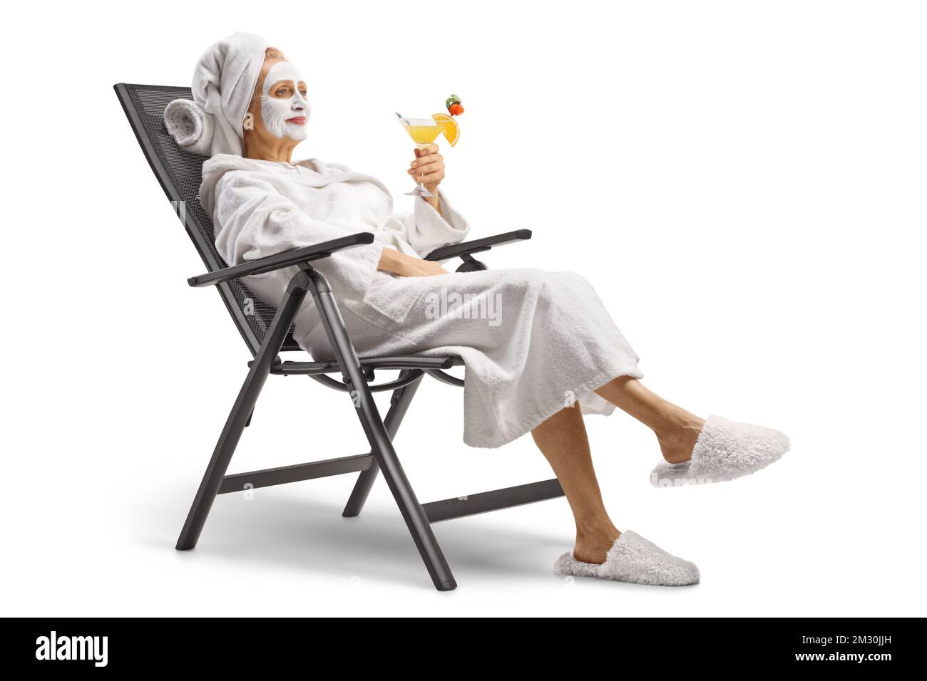 Donna in accappatoio con maschera facciale seduta su una sedia e rilassante con un cocktail isolato su sfondo bianco Foto Stock