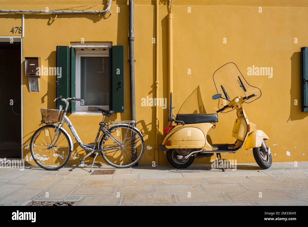 Street color Italia, vista in estate di uno scooter Vespa e di una bicicletta parcheggiata contro un muro giallo nella città veneziana di Chioggia, Veneto, Italia, Foto Stock