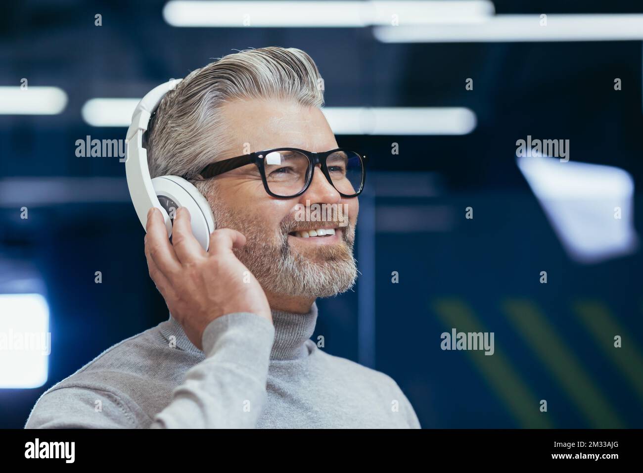 Primo piano. Ritratto di un vecchio uomo dai capelli grigi in occhiali che ascolta musica in cuffie in ufficio, tenendo le cuffie in mano, sorridendo, guardando lontano. Foto Stock