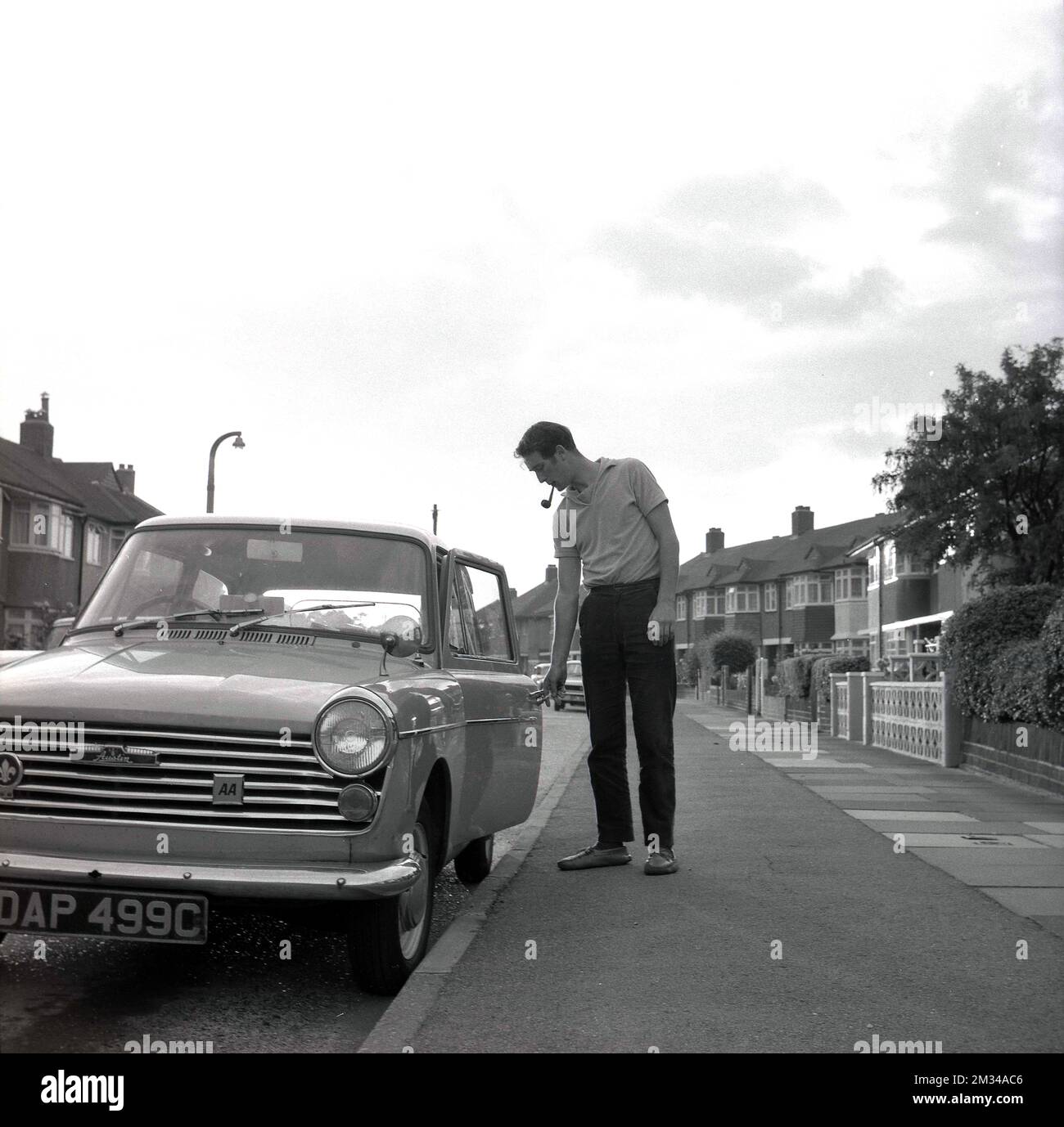 1960s, storico, un uomo in piedi fuori sul marciapiede in una strada suburbana che apre la porta della sua auto, una Austin A40 farina, una piccola economia auto dell'epoca, che era in produzione tra il 1958 e il 1967. Il suo nome deriva dal designer dell'auto, lo studio 'Pinin farina' di Torino. Foto Stock