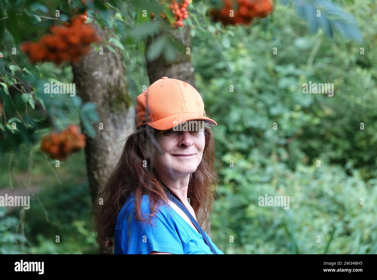 Donna anziana con cappuccio arancione sotto i grappoli di bacche d'arancia sfocati di un albero di rowan Foto Stock
