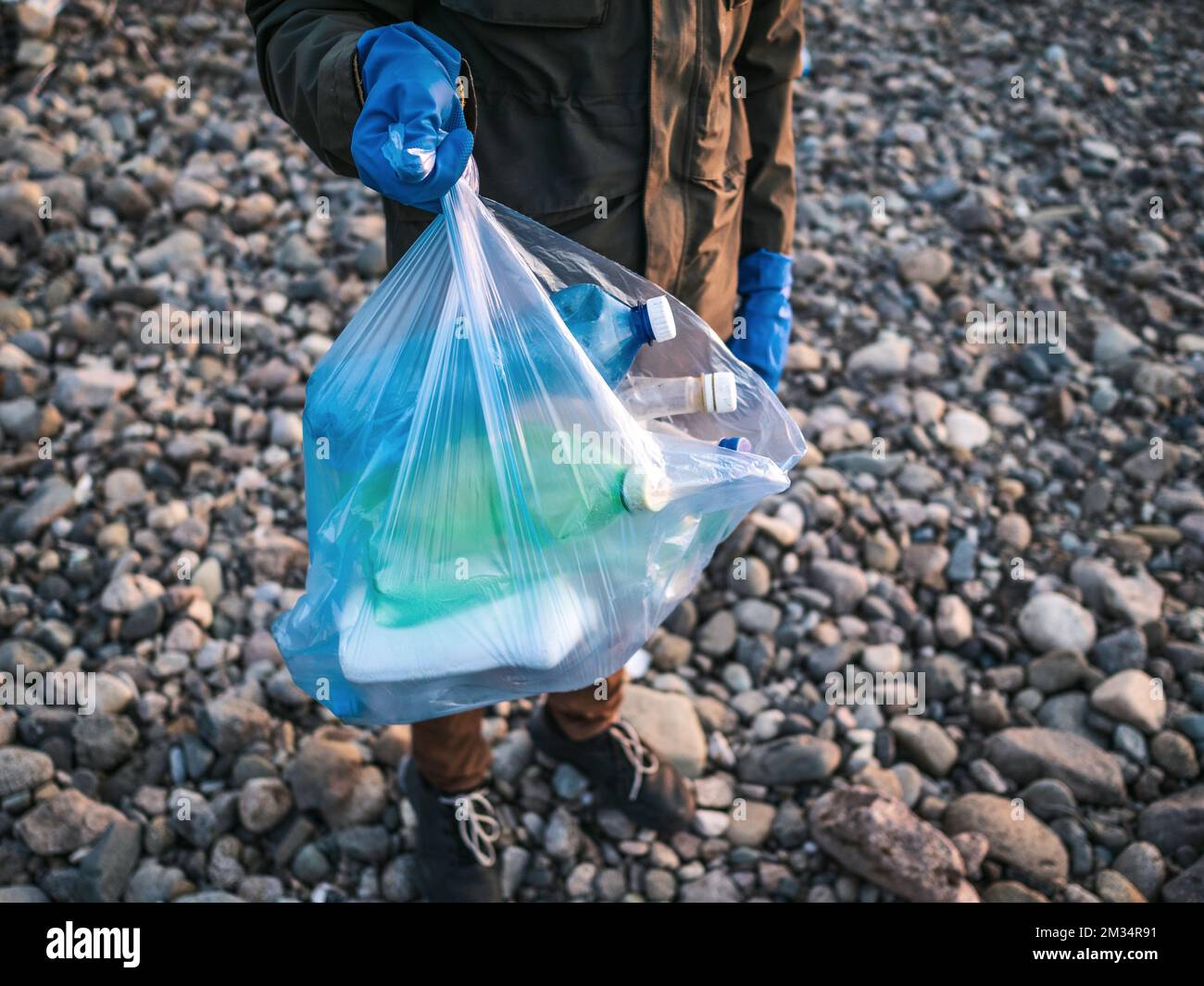 Processo di pulizia della spiaggia di pietra dai rifiuti di plastica. L'uomo tiene bottiglie di plastica nel sacchetto dei rifiuti. Concetto di inquinamento ambientale. Foto Stock