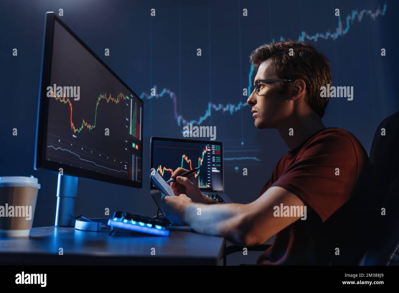 Vista laterale di un analista professionista giovane al tavolo di lavoro, analisi del grafico finanziario, quotazioni azionarie, prezzi delle azioni, trading online, verifica dei dati sul grafico della criptovaluta sullo schermo del computer, prendendo appunti Foto Stock