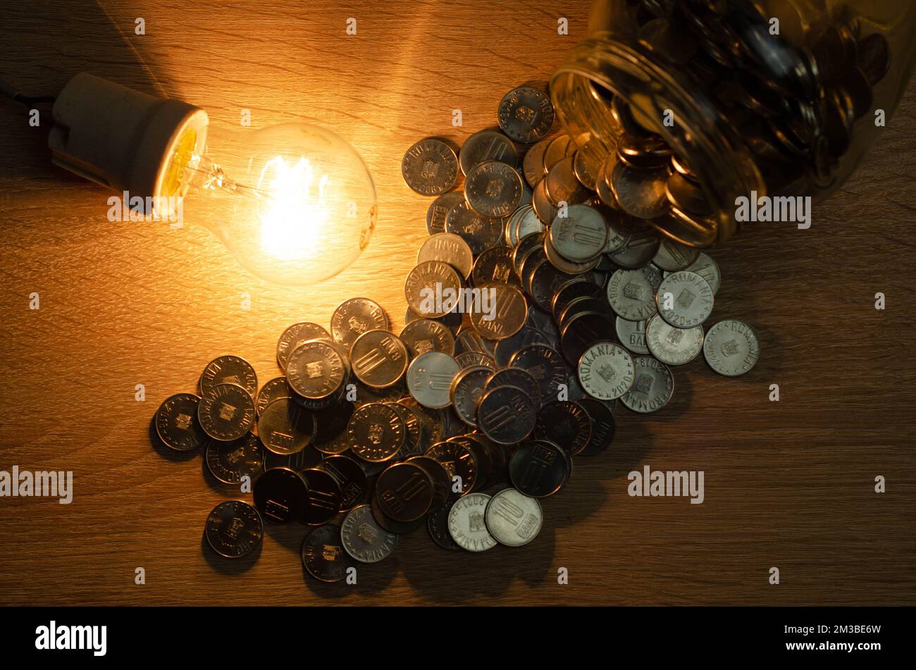 Concetto di consumo di elettricità, in questi tempi di crisi, rappresentato da una lampadina e monete in euro sparse, sfondo scuro Foto Stock