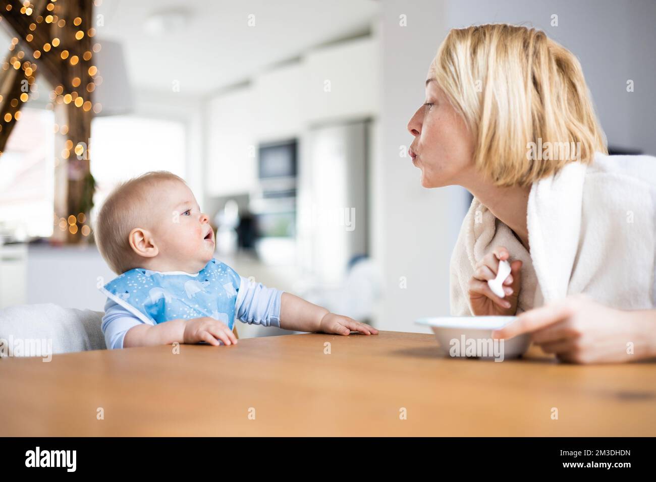 Madre che motiva il bambino mentre lo nutre seduto in poltrona al tavolo da pranzo a casa. Concetto di introduzione degli alimenti solidi per bambini. Foto Stock