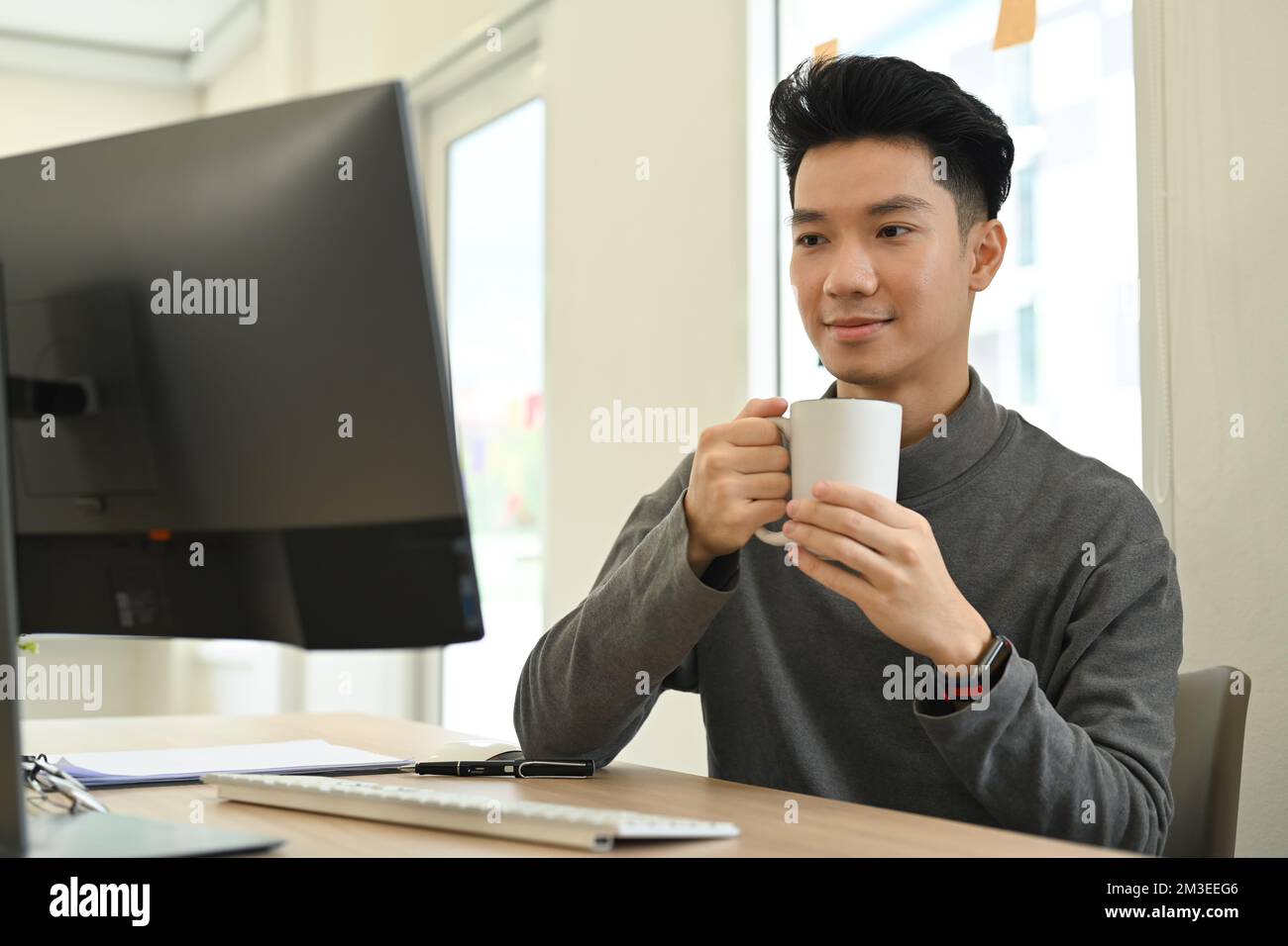Uomo asiatico sorridente che tiene una tazza di caffè e guarda il webinar online sullo schermo del computer Foto Stock