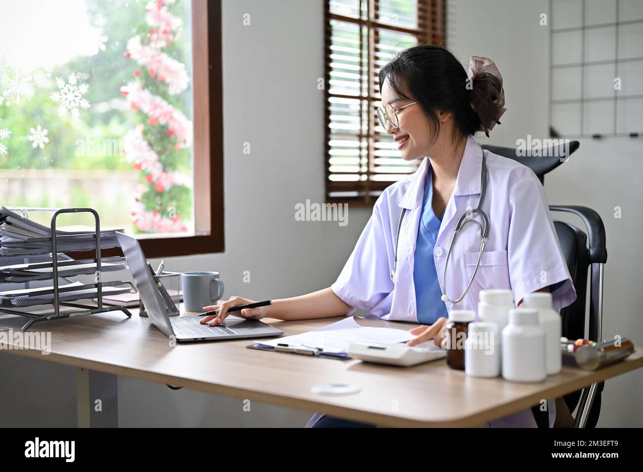 Attraente medico asiatico utilizzando calcolatrice e computer portatile, stimare il costo della chirurgia per la sua paziente, lavorando in ufficio. Foto Stock