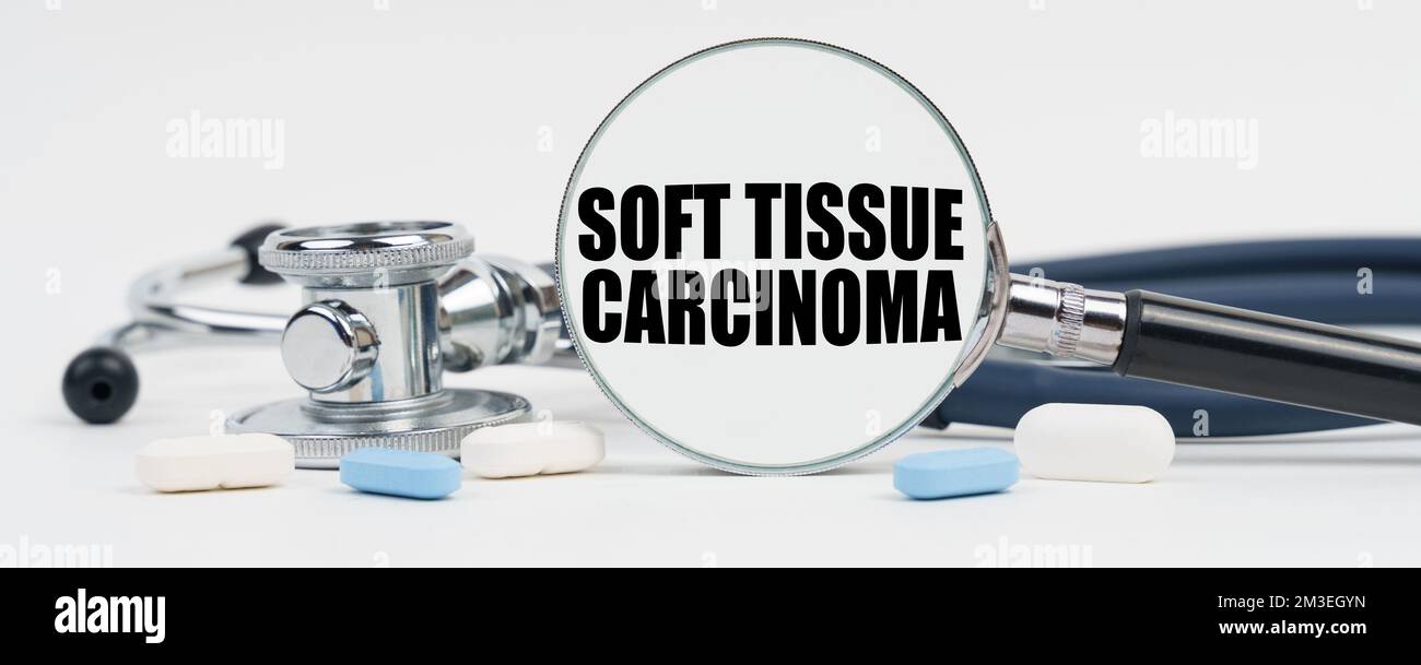 Medicina e concetto di salute. Su una superficie bianca ci sono pillole, uno stetoscopio e una lente d'ingrandimento al cui interno è scritto - carcinoma del tessuto molle Foto Stock