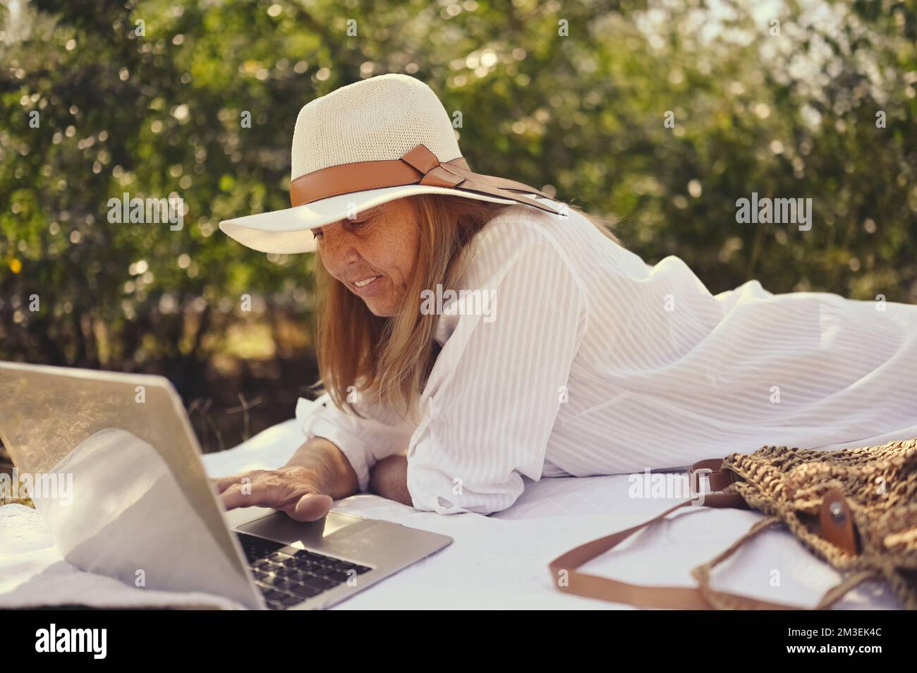 Donna anziana felice che lavora o videochiama con un computer portatile in estate all'aperto. Vecchia ed elegante signora in cappello di paglia e borsa seduta sull'erba durante il picnic in campagna. Concetto attivo delle persone in pensione Foto Stock