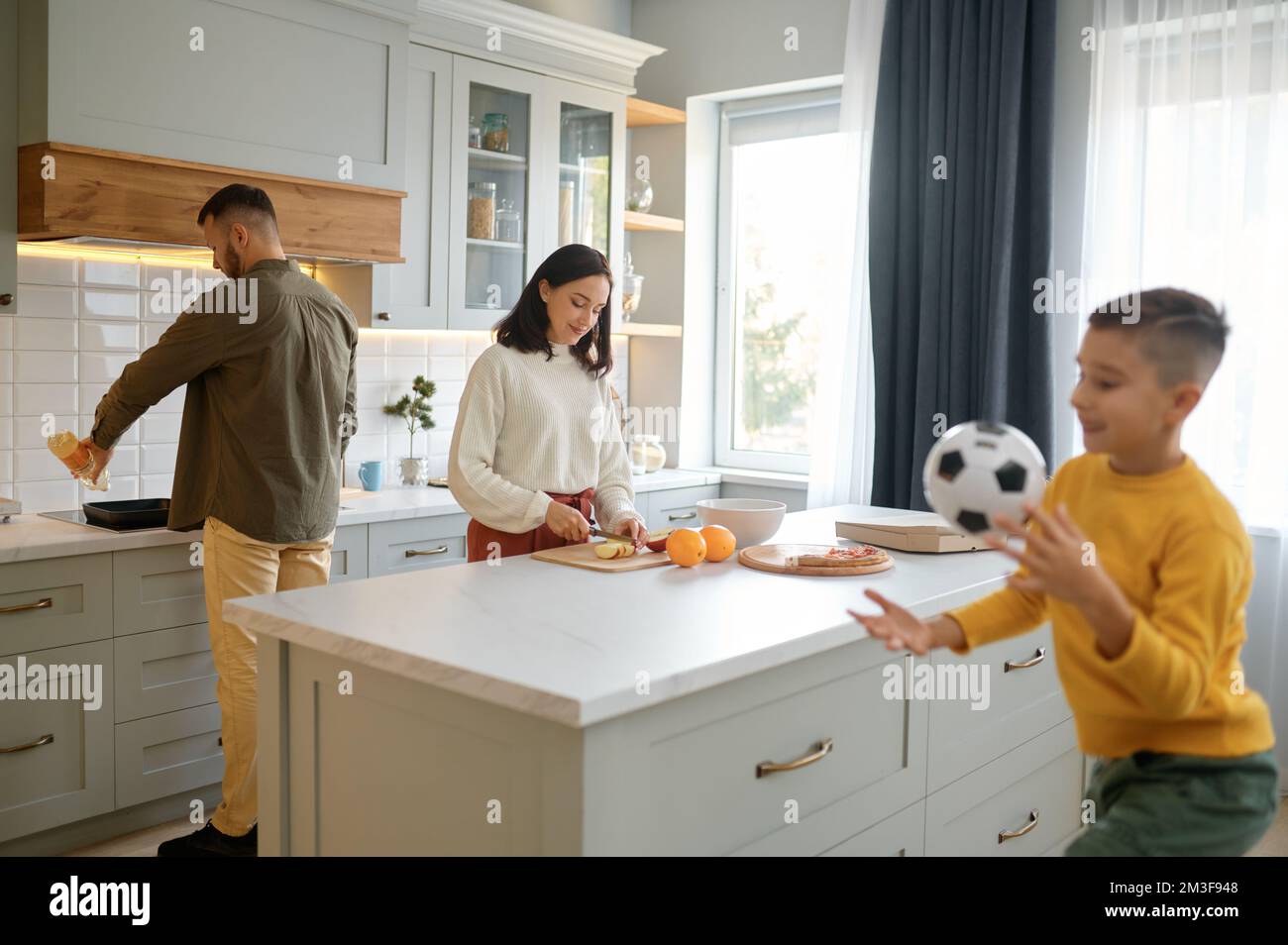 I genitori cucinano in cucina mentre il figlio del ragazzo gioca la sfera di calcio Foto Stock