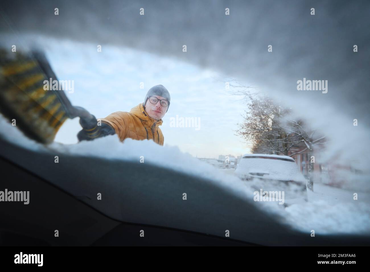 Uomo che sgomberava la neve dal parabrezza la sua auto innevata dopo la caduta di neve. Parcheggio in strada in inverno. Foto Stock