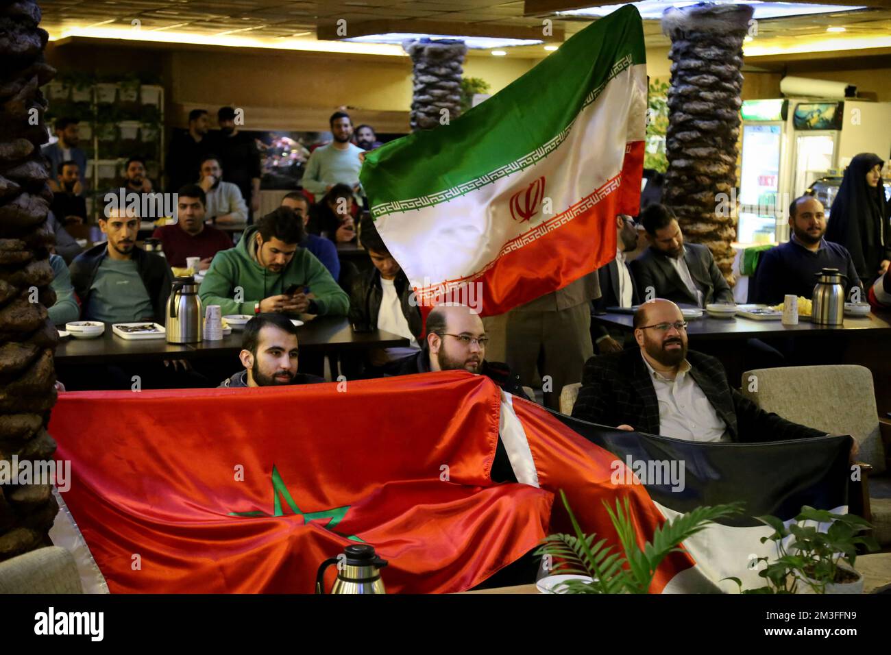 Teheran, Teheran, Iran. 14th Dec, 2022. Gli uomini iraniani detengono una bandiera palestinese (R) e una bandiera marocchina (L) e una bandiera iraniana (TOP) mentre un gruppo di tifosi palestinesi seduti dietro di loro durante la Coppa del mondo FIFA Qatar 2022 partita semifinale tra Francia e Marocco, al caffè Nakhlestan nel centro di Teheran, il 14 dicembre 2022. Tifosi provenienti da Iran, Libano, Yemen, Siria e Palestina si riuniscono al caffè Nakhlestan, gestito e di proprietà dell'Owj, Islamic Revolutionary Guard Corps (IRGS) Arts and Media Organization, per sostenere la nazionale marocchina. Dopo la vittoria contro il Portogallo in Qatar o Foto Stock