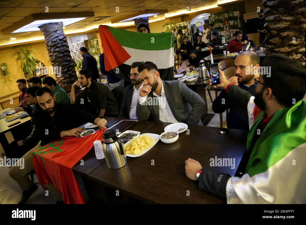 Teheran, Teheran, Iran. 14th Dec, 2022. Un uomo iraniano detiene una bandiera palestinese mentre un gruppo di tifosi palestinesi seduti dietro di loro durante la Coppa del mondo FIFA Qatar 2022 partita semifinale tra Francia e Marocco, al caffè Nakhlestan nel centro di Teheran, il 14 dicembre 2022. Tifosi provenienti da Iran, Libano, Yemen, Siria e Palestina si riuniscono al caffè Nakhlestan, gestito e di proprietà dell'Owj, Islamic Revolutionary Guard Corps (IRGS) Arts and Media Organization, per sostenere la nazionale marocchina. Dopo la vittoria contro il Portogallo in Qatar il 10th dicembre, i marocchini portavano bandiere palestinesi Foto Stock