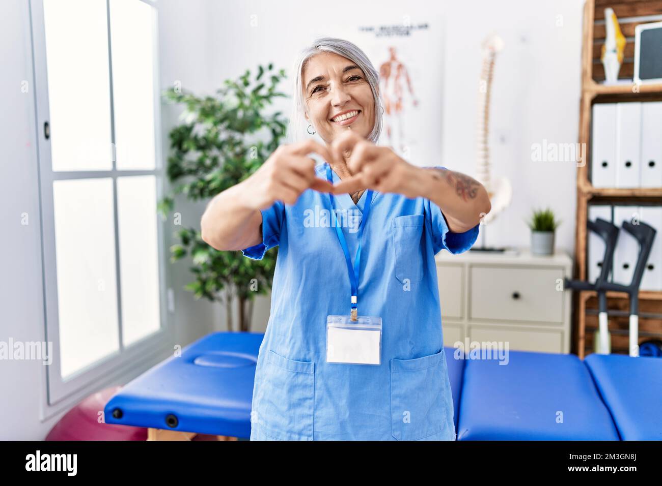 Donna di mezza età dai capelli grigi che indossa l'uniforme fisioterapista alla clinica medica sorridente nell'amore che fa la forma del simbolo del cuore con le mani. concetto romantico. Foto Stock