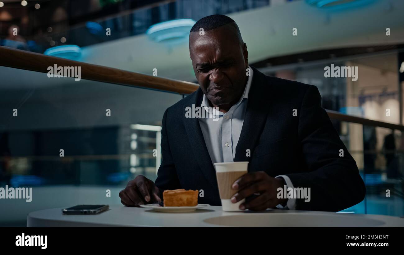 Uomo d'affari afro-americano di mezza età imprenditore caffè ospite uomo seduto mangiare in caffetteria insoddisfatto con torta cattiva dissotenza torta dessert Foto Stock