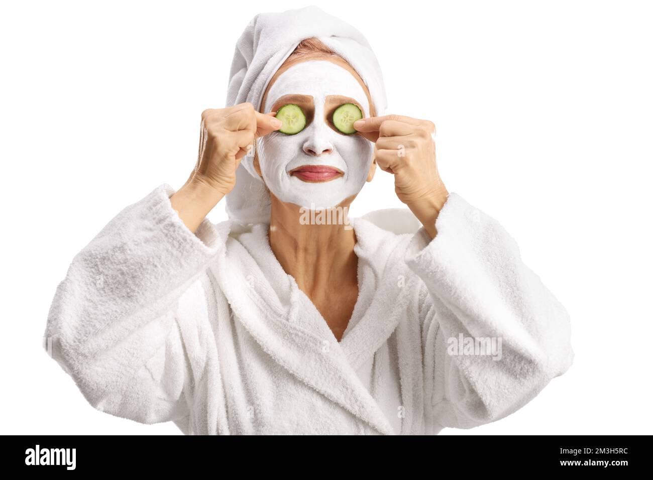 Donna in un accappatoio bianco con una maschera viso che mette su cetrioli sugli occhi isolati su sfondo bianco Foto Stock