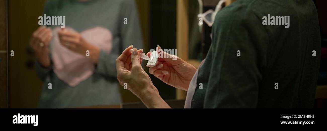 Immagine a vista ampia delle mani femminili che gocciolano acqua di analisi salina su una penna per test del kit home test per coronavirus. Con un riflesso nello specchio del bagno. Foto Stock