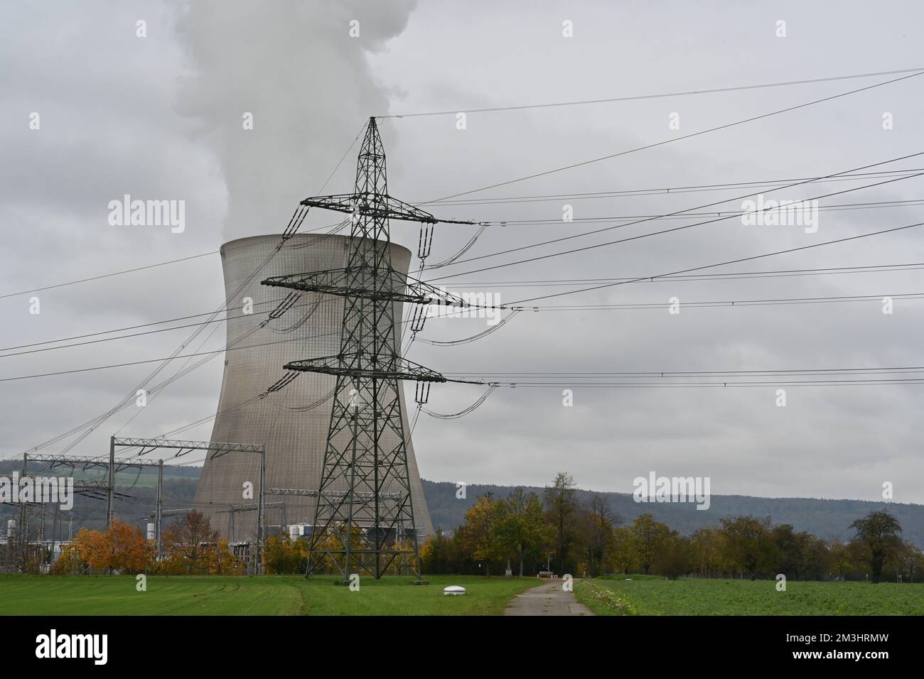 Vista sul pilastro elettrico di fronte alla torre di raffreddamento di una centrale nucleare con fumo sotto le nuvole. Foto Stock