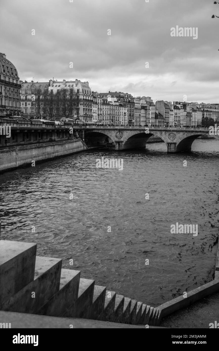 Scala di grigi verticale di un vecchio ponte su un fiume che scorre con un paesaggio urbano sullo sfondo Foto Stock