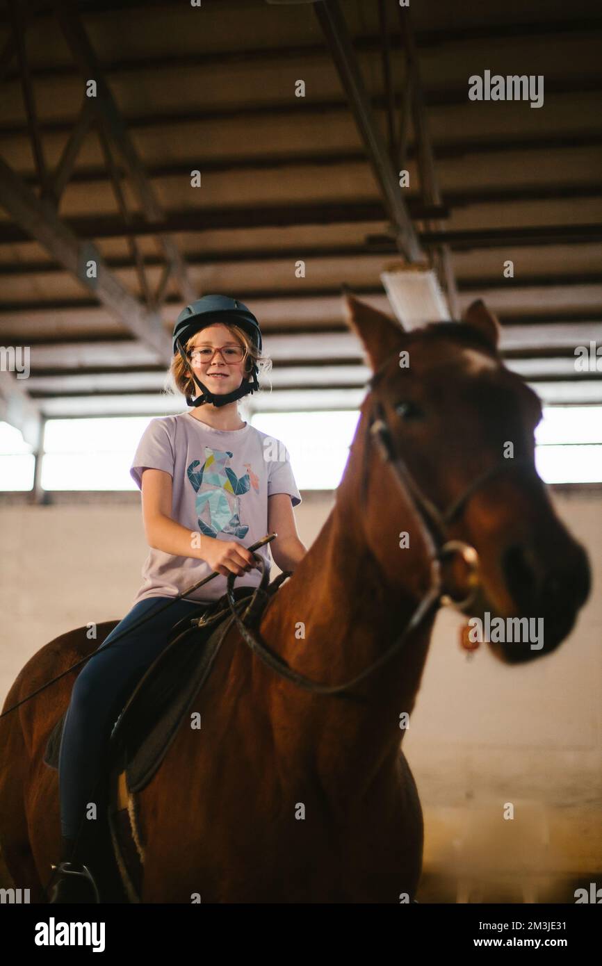 Ragazza in bicchieri sorride a cavallo nella lezione di equitazione in fienile Foto Stock