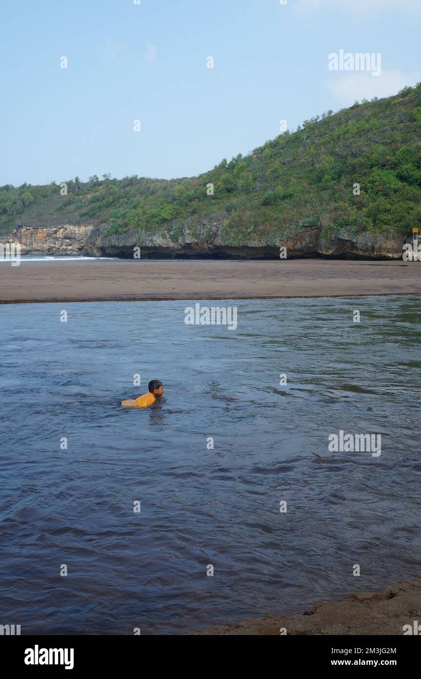 Bellissima spiaggia di mare circondata da colline rocciose con cielo blu. Un ragazzo asiatico che nuota in acqua Foto Stock