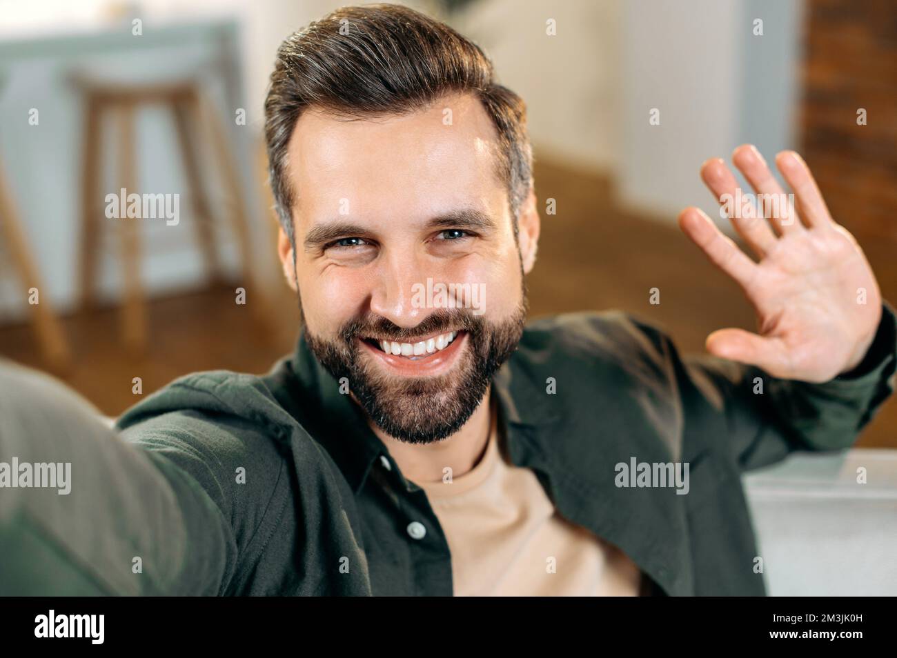 Allegro attraente positivo uomo caucasico bearded, in una camicia casual, prende un selfie sul suo smartphone, le onde mano, guarda nella fotocamera anteriore del telefono cellulare, sorrisi amichevole Foto Stock