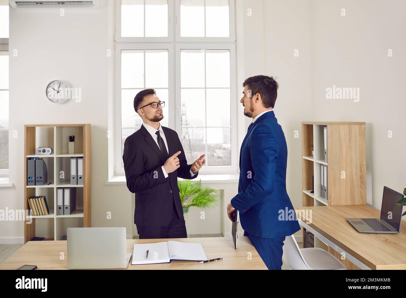 Due giovani uomini d'affari che hanno una conversazione mentre sono in piedi accanto a una scrivania dell'ufficio Foto Stock