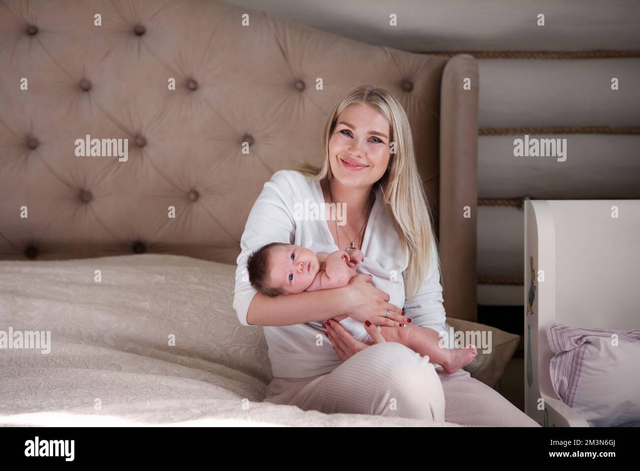 La madre felice tiene il bambino neonato fra le braccia, sorride seduto sul letto e guarda la macchina fotografica. Bella mamma bionda con capelli lunghi e suo bambino a casa Foto Stock