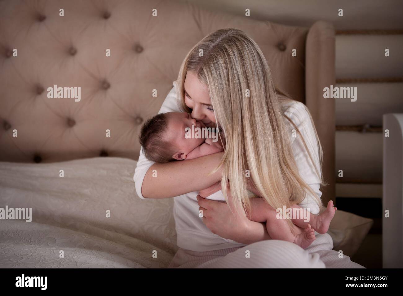 La madre bacia il neonato tra le braccia, seduto a letto. Bella mamma bionda con capelli lunghi e suo bambino a casa. Amore e tenerezza Foto Stock