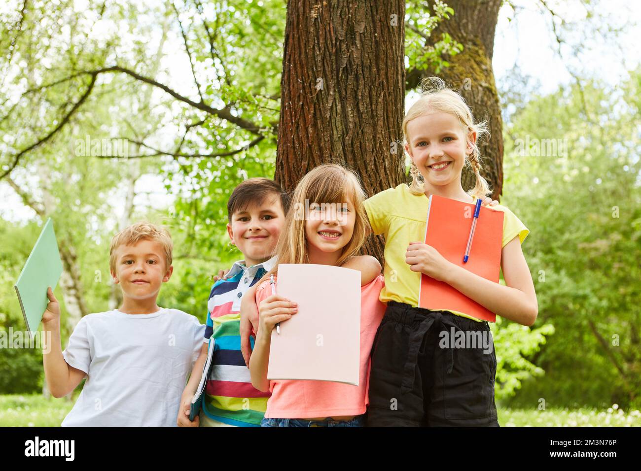 Ritratto di amici maschi e femmine sorridenti braccia intorno tenendo libri contro l'albero in giardino Foto Stock
