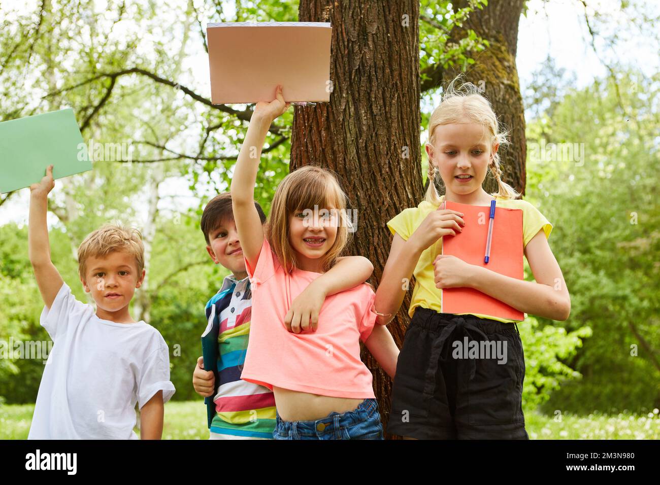 Ragazzi e ragazze multirazziali che tengono libri mentre si trovano contro l'albero nel parco durante le vacanze estive Foto Stock