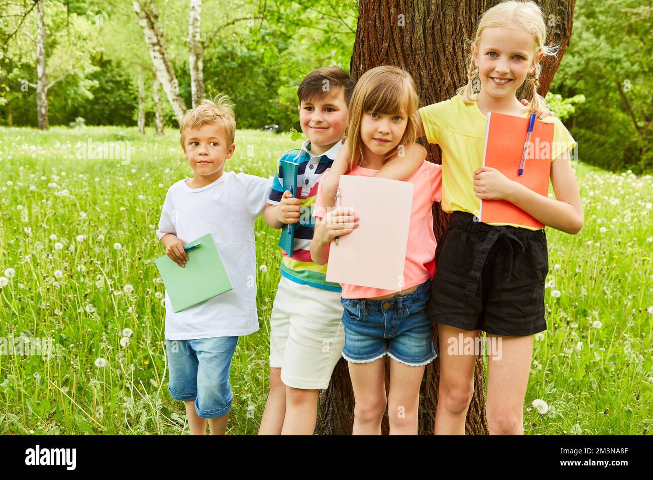 Ritratto di amici maschi e femmine braccia intorno tenendo libri contro l'albero in giardino Foto Stock