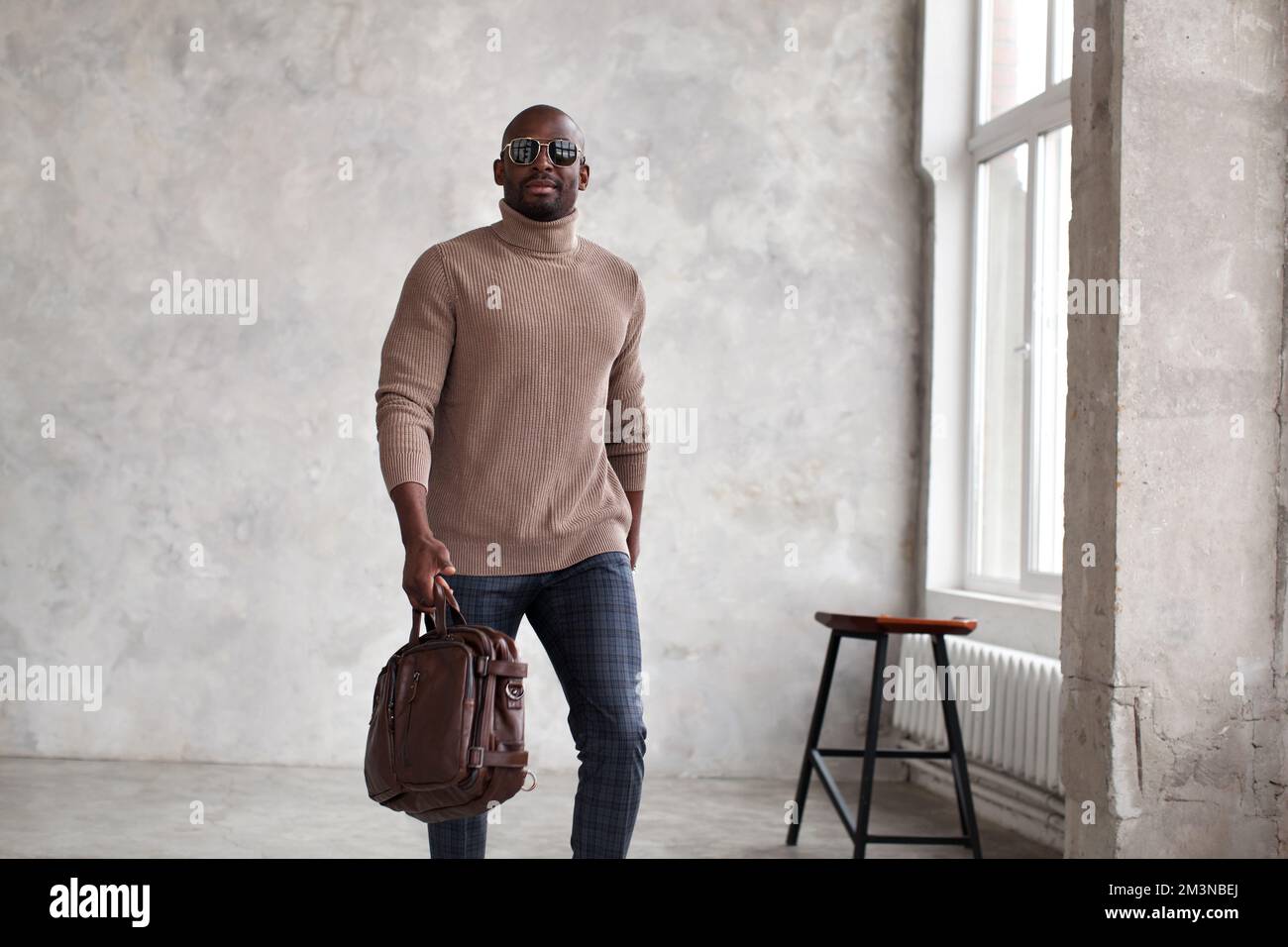 Bell'uomo afroamericano che cammina all'interno indossando abiti alla moda, maglione beige, pantaloni e borsa in pelle, occhiali da sole Foto Stock