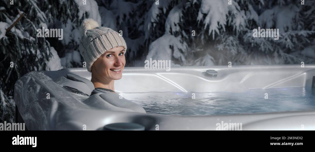 donna sorridente con cappello lavorato a maglia che si rilassa nella vasca idromassaggio all'aperto durante l'inverno innevato. banner con spazio di copia Foto Stock