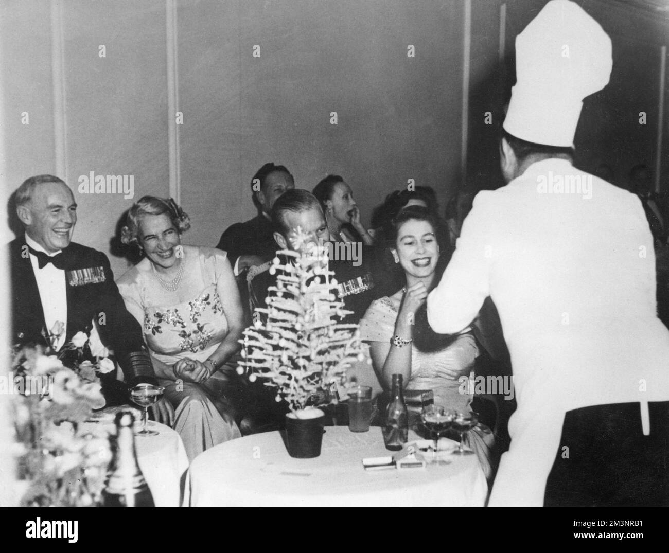 La principessa Elisabetta (Regina Elisabetta II) partecipa a una danza tenuta all'Hotel Phoenicia a Malta dalla Sala di guerra Mess of Naval Headquarters, Lascaris. Una caratteristica speciale era un cabaret di ufficiali e qui la principessa sta sorridendo acquistando un hot dog dal capitano ben Fisher, capitano della flotta, vestito da chef. Il Duca di Edimburgo è in parte nascosto dietro un albero di Natale in miniatura, e sulla sinistra sono l'ammiraglio Sir John Edelsten, Comandante-in-Capo della flotta Mediterranea e Lady Edelsten. Data: 1951 Foto Stock