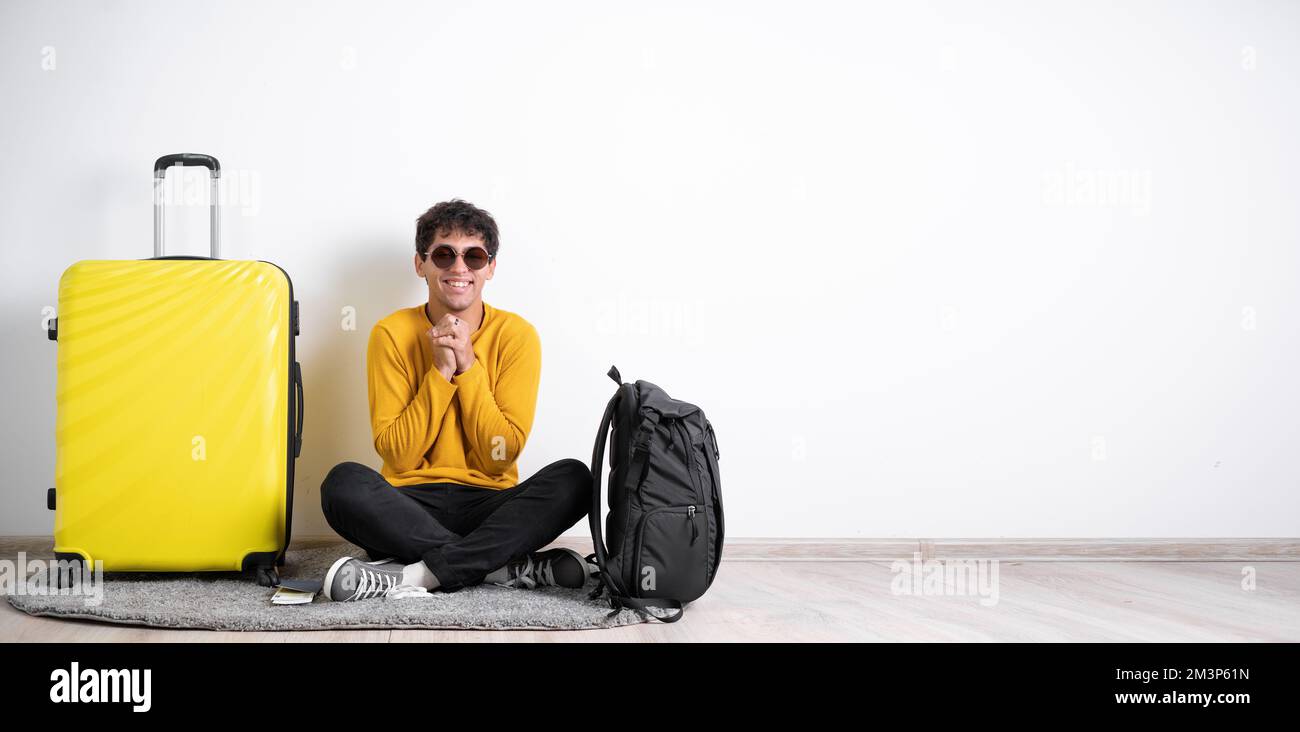 Il giovane viaggiatore con una valigia soddisfatto di un'espressione positiva si siede isolato su uno sfondo bianco tiene le mani vicino al petto Foto Stock