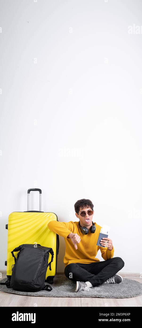 Giovane uomo arabo misto corsa infelice con valigia e documento pronto per andare a viaggiare mostrando pollice giù ed esprimendo antipatico. Banner verticale Foto Stock