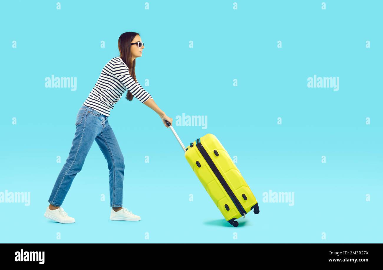 Vista laterale di una giovane donna con valigia da viaggio gialla che cammina su sfondo blu Foto Stock