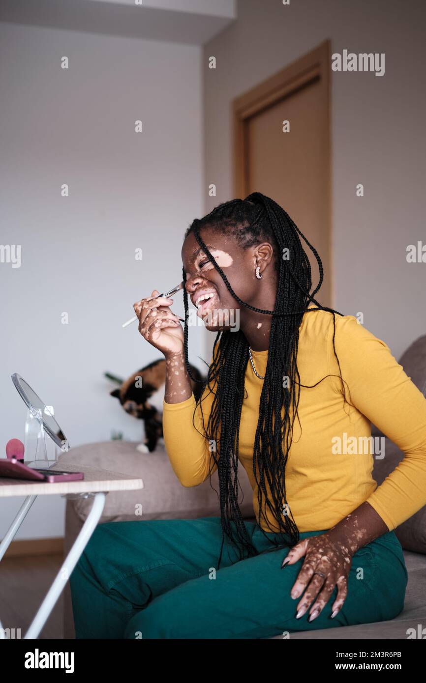 Donna afro-americana felice con le macchie della pelle seduta sul divano che fa il trucco Foto Stock
