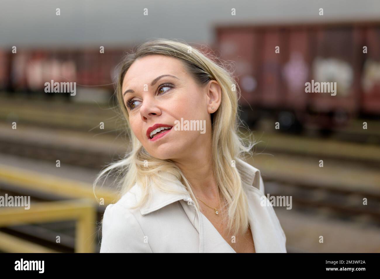 Primo piano di un'attraente donna bionda di mezza età in un camice bianco si erge davanti alle carrozze ferroviarie e guarda sopra la spalla con un cappotto Foto Stock