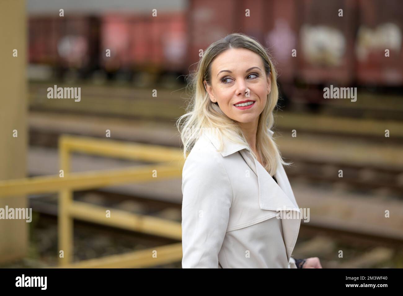 Bella donna bionda di mezza età in un cappotto di trincea bianco si trova davanti alle carrozze ferroviarie e guarda sopra la sua spalla con un felice ridere Foto Stock