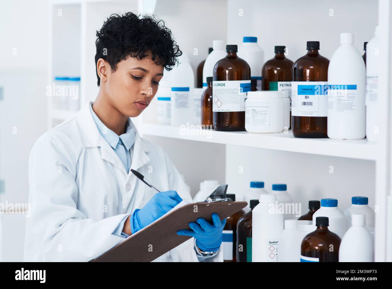 Annotando tutte le scorte che ha nel suo laboratorio. un giovane scienziato che scrive su una clipboard accanto a uno scaffale di sostanze chimiche in un laboratorio. Foto Stock