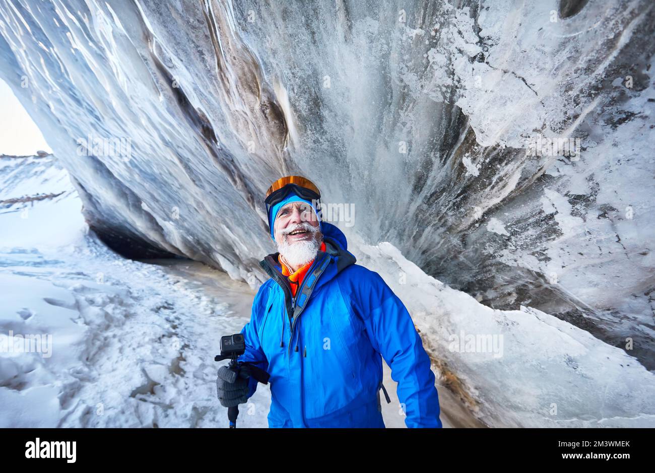 Ritratto di un vecchio uomo sorridente portatore con Action camera in costume blu vicino alla parete di una grotta di ghiaccio sul ghiacciaio nella valle di montagna coperta di neve ad Almaty, K Foto Stock