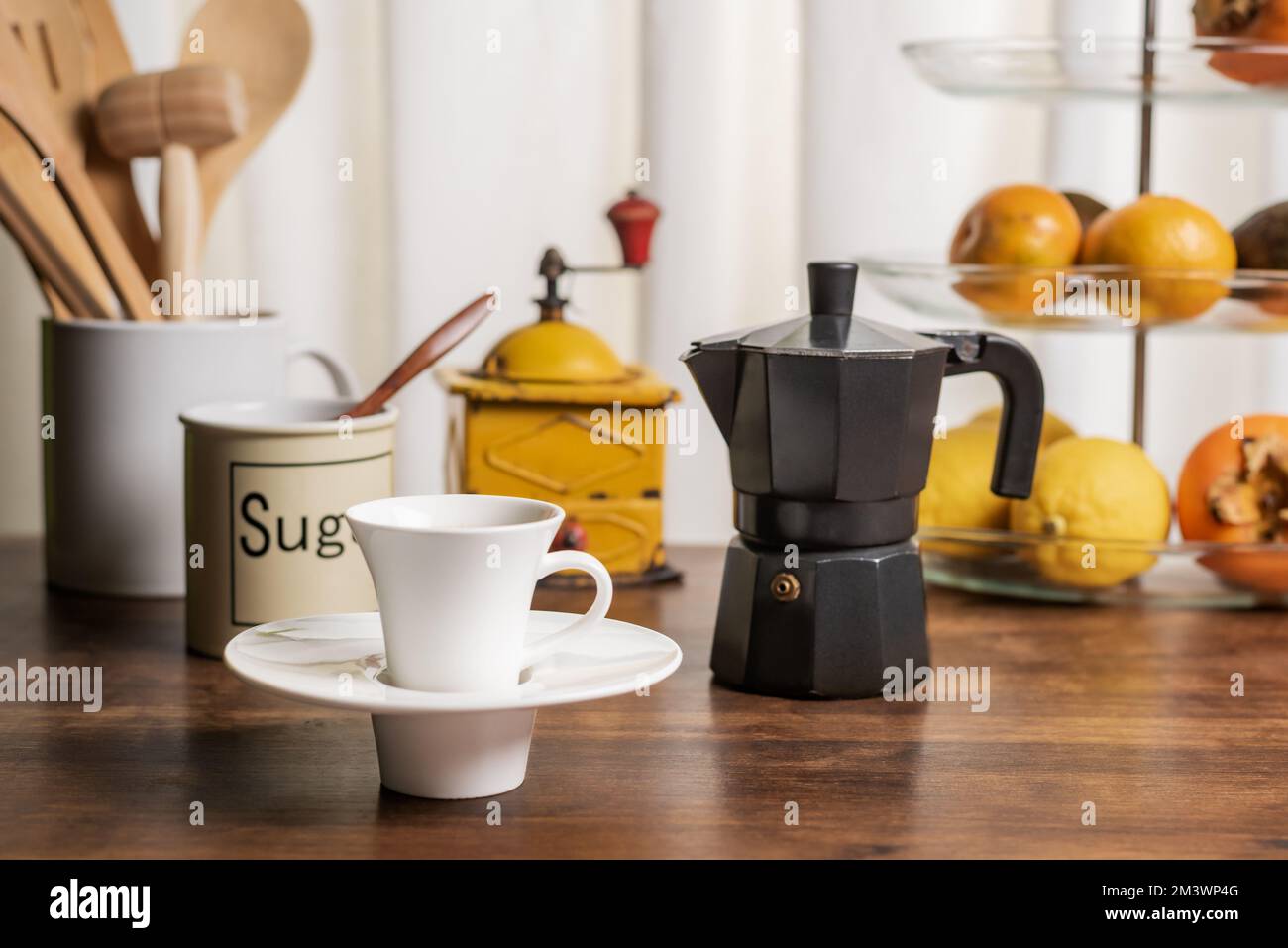 Tazza di caffè su un tavolo da cucina con un recipiente di frutta, una caraffa con utensili in legno, un macinacaffè e una macchina per il caffè italiana. Foto Stock