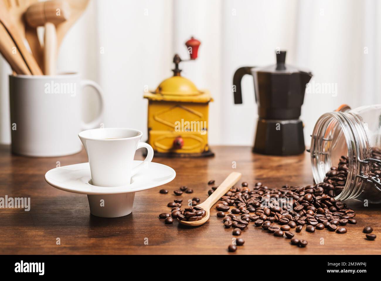 Tazza di caffè su un tavolo da cucina con una caraffa con utensili in legno, un macinacaffè, una macchina per il caffè italiano e un vaso rovesciato con caffè tostato Foto Stock